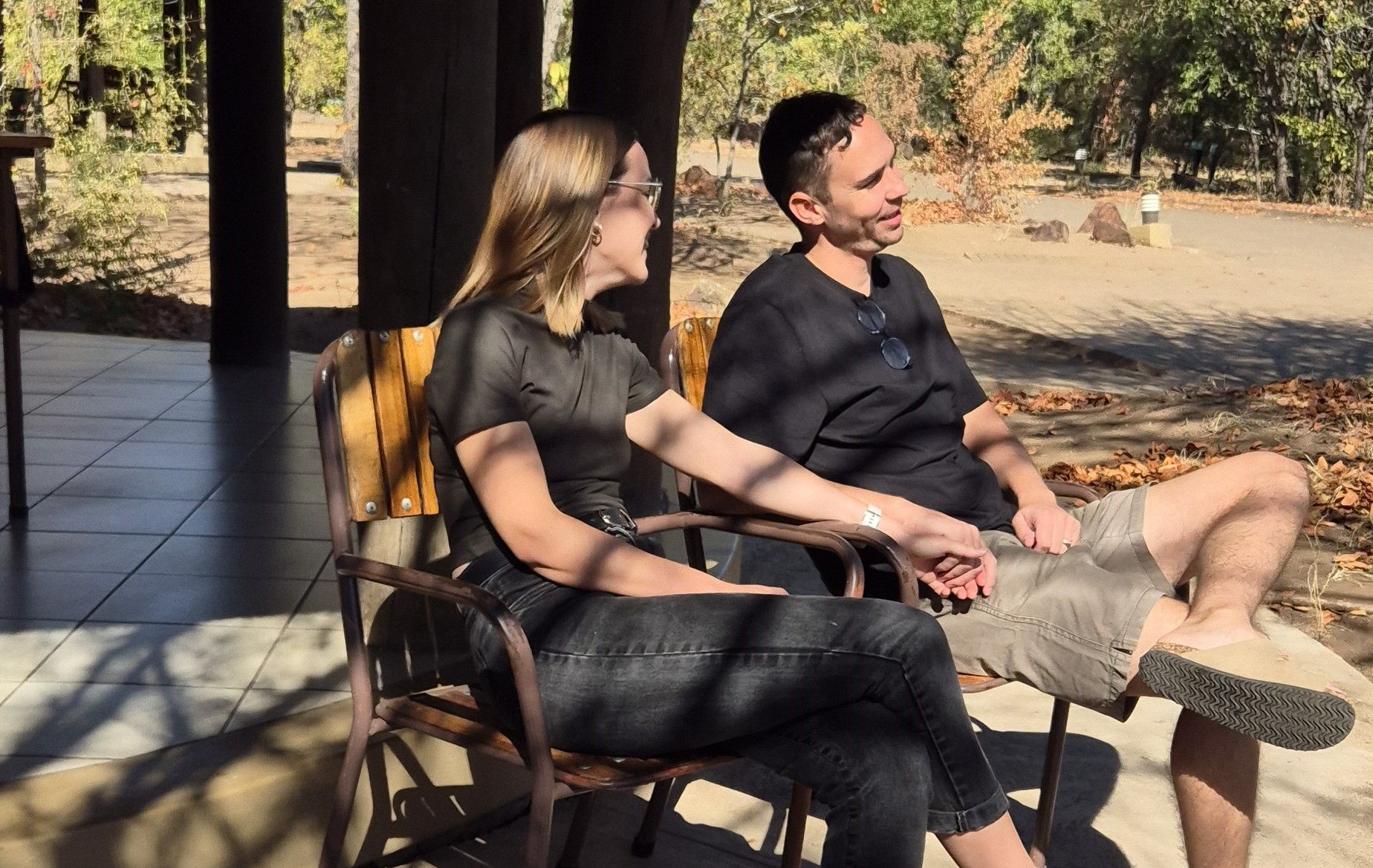 Two people sit outdoors on chairs, smiling and chatting in the sun.