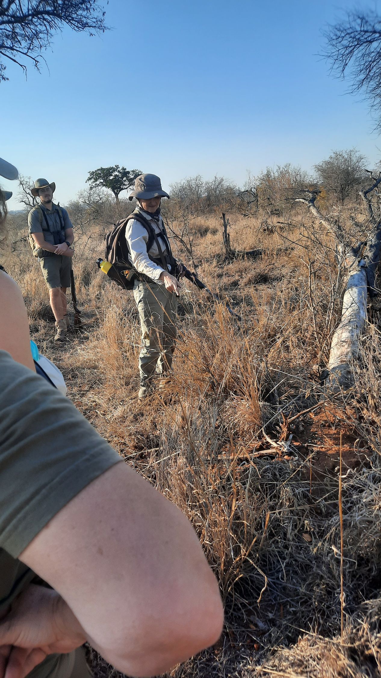 People with gear in a dry, brown landscape; one holds a rifle, appears to be pointing.