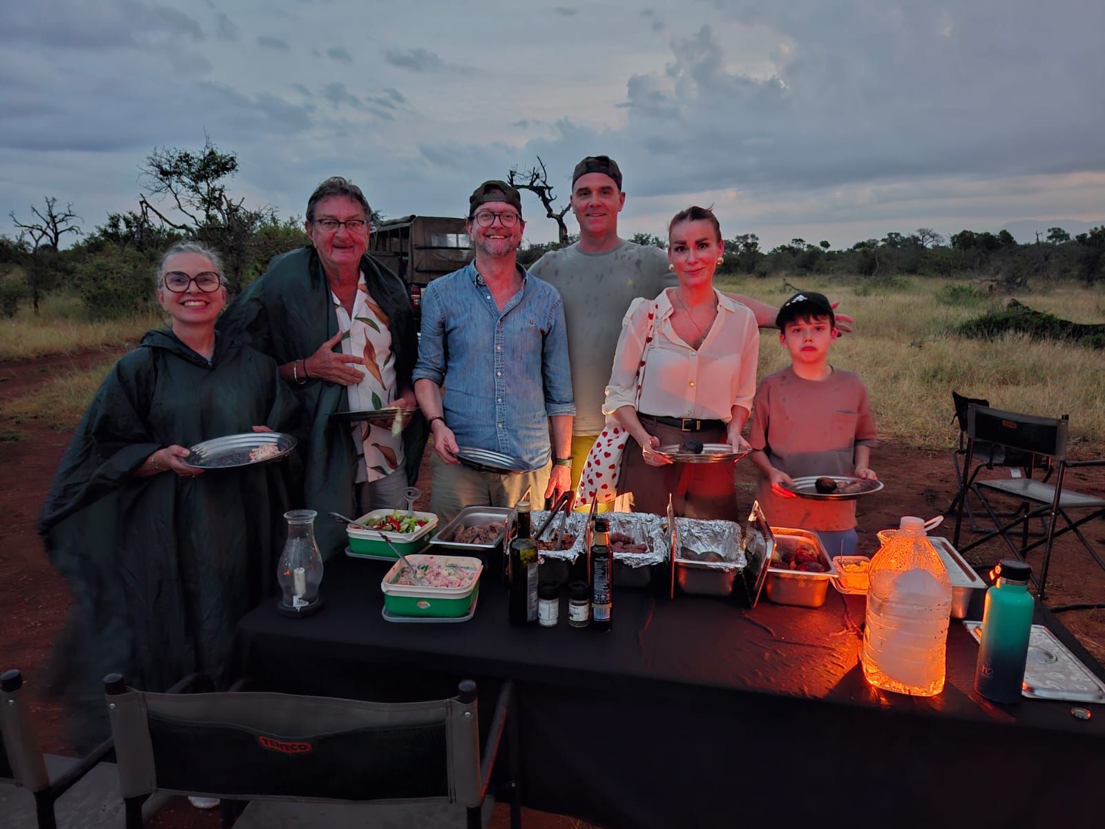 Group gathered outdoors at dusk behind a table with food and drinks, smiling in a grassy field.