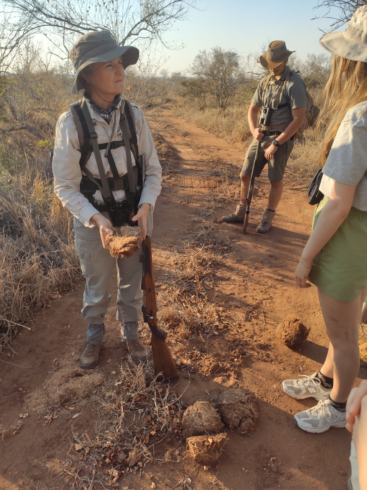 A person holding dung, explaining to others on a dusty path in a sunny savanna.
