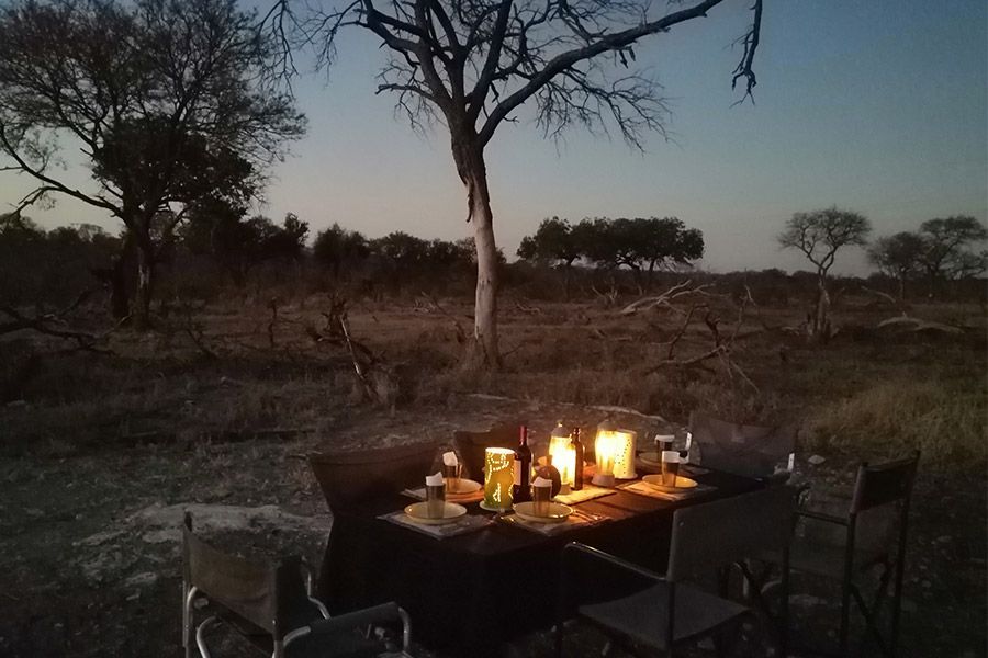 A candlelit dinner table set for two outdoors at dusk, surrounded by trees in a dry, open savannah landscape.