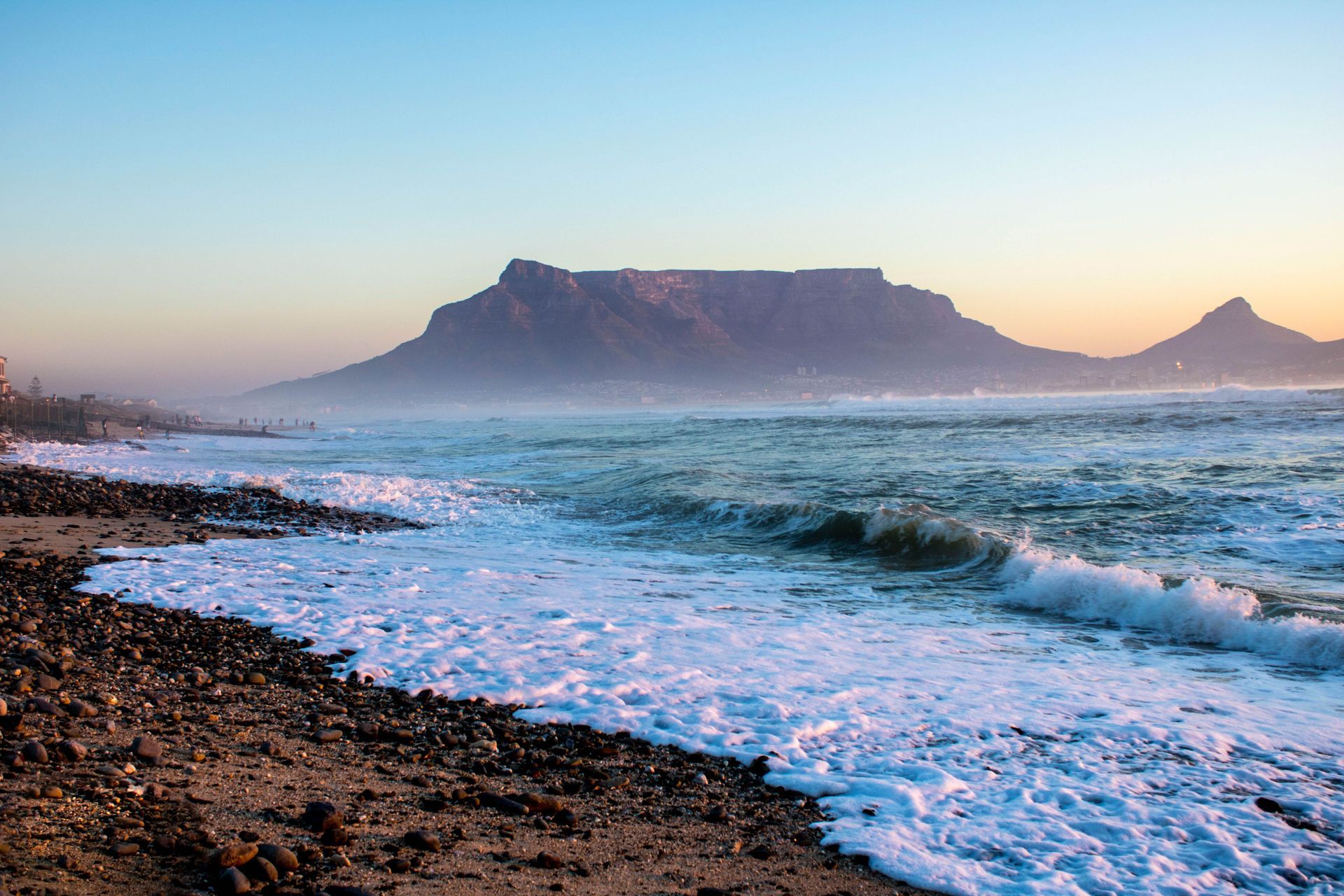 Ocean waves in the foreground with Table Mountain at sunrise and a rocky shoreline.