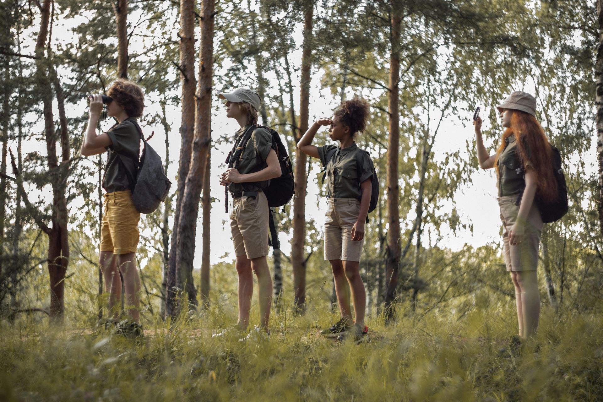 Four children exploring a forest; one uses binoculars, others point. They wear hats and khaki shorts.
