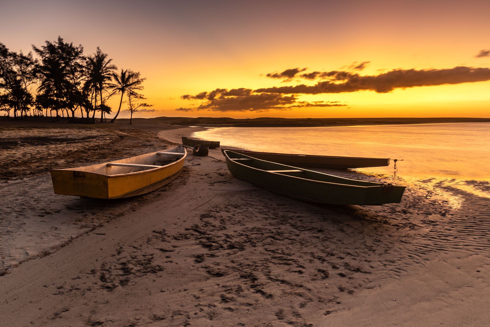 Two boats on a sandy beach at sunset, with palm trees silhouetted along the shore.