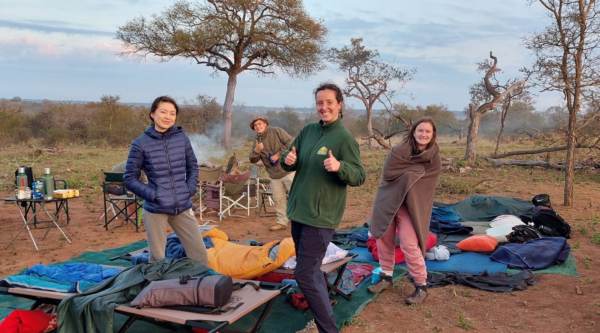 Three people smiling by camping gear at sunset in a dry, open field with sparse trees.