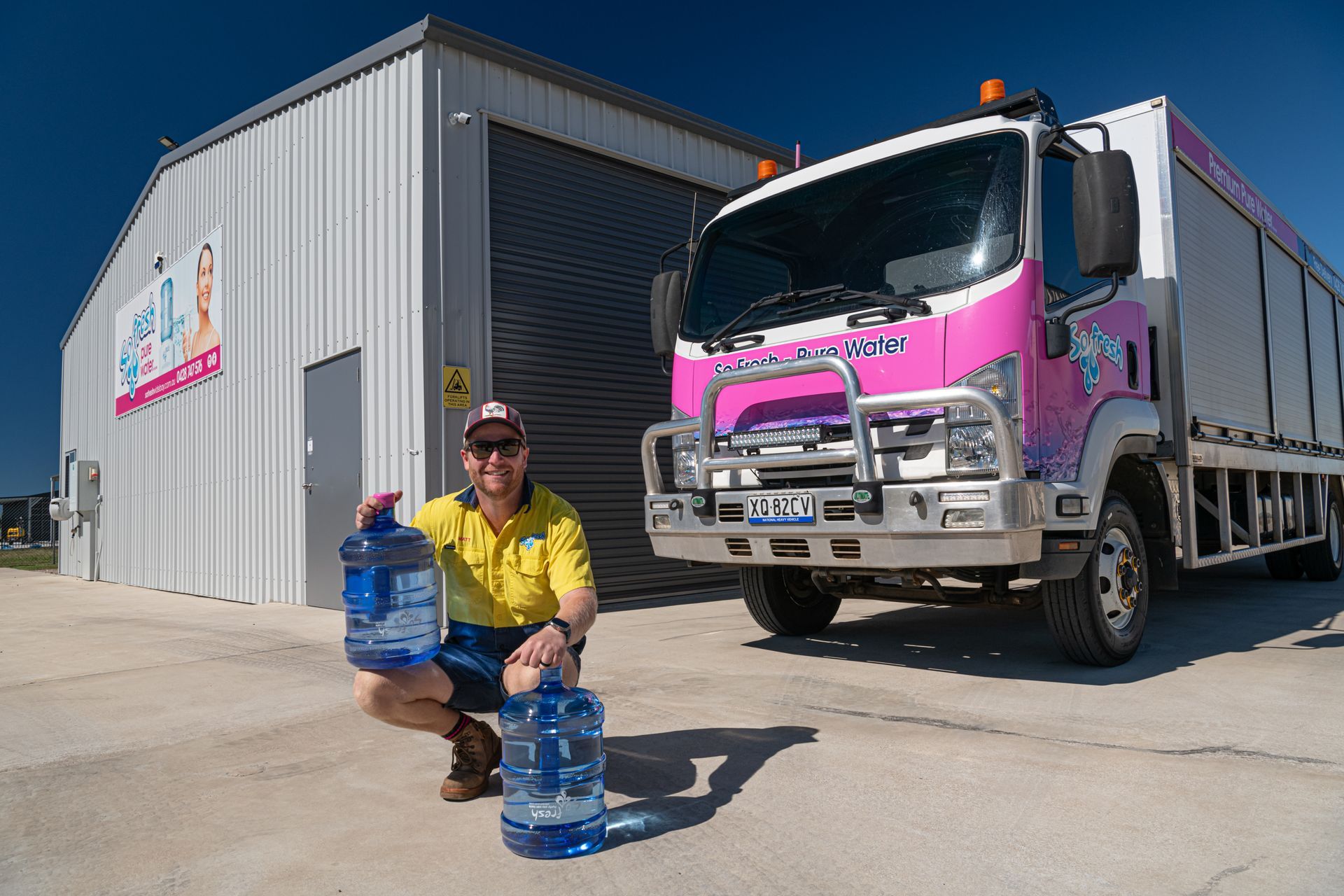 Worker crouches by water jugs beside a pink truck outside a warehouse — So Fresh Wide Bay in Thabeban, QLD