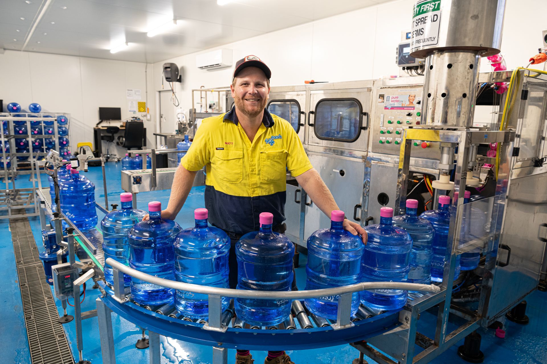 Worker in yellow shirt standing by blue water jugs on a bottling line in a factory — So Fresh Wide Bay in Thabeban, QLD