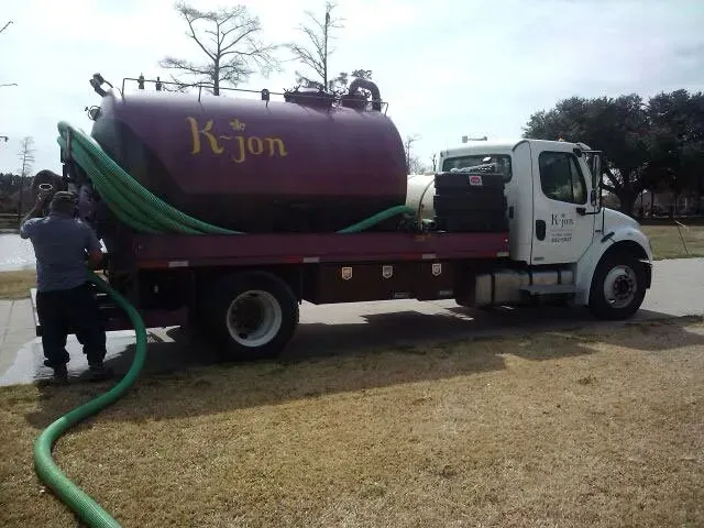 Septic tank truck with worker connecting a green hose on a residential lawn.