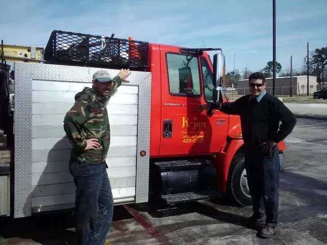 Two men standing next to a red work truck with silver door. One in camo, other in black sweater. Sunny day.