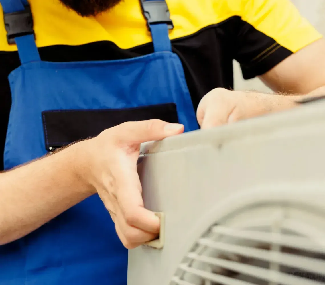 A person in a yellow hard hat and blue overalls works on the metal panel of an outdoor air conditioning unit.