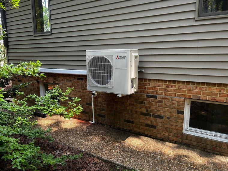 A gray American Standard air conditioning unit sitting on a concrete pad next to a house with beige siding.