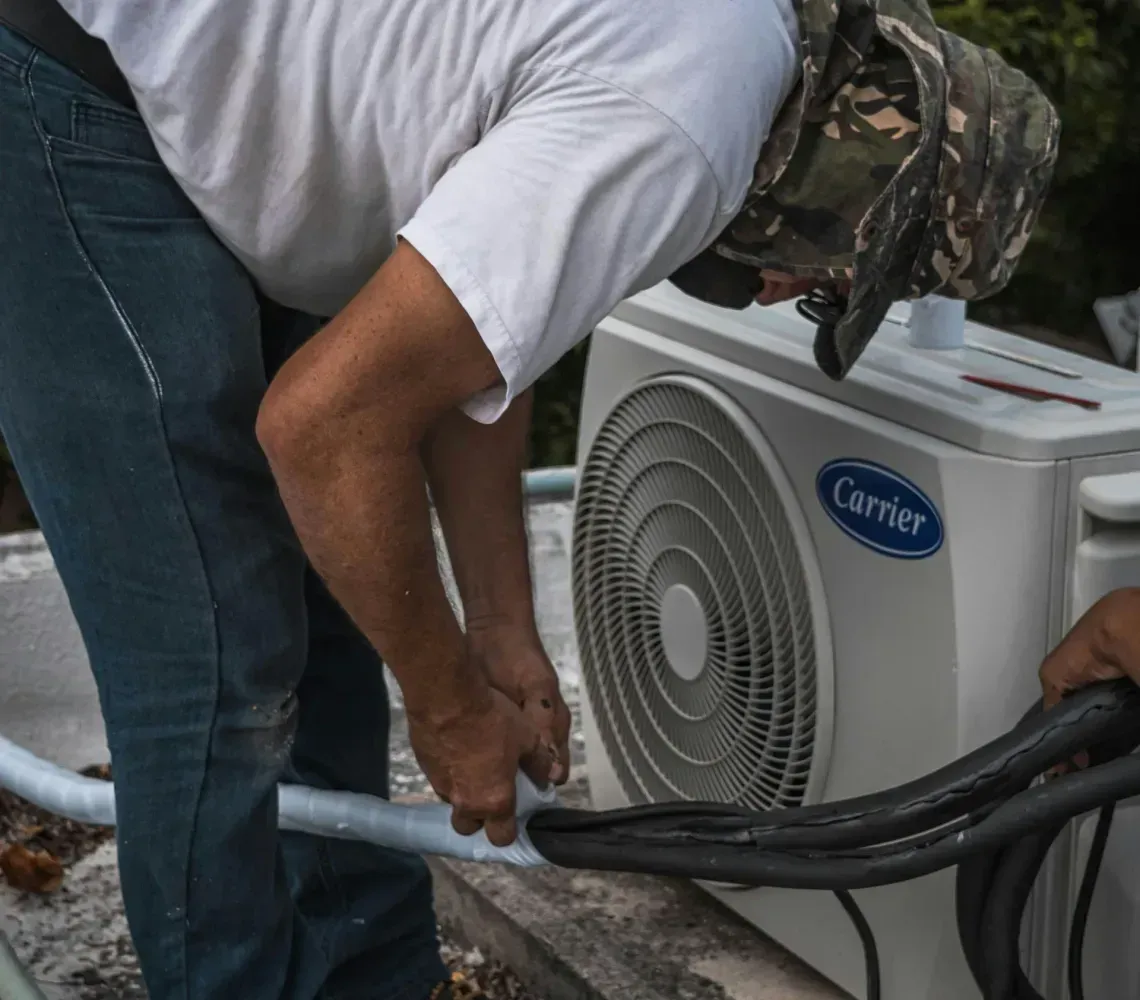 A technician in work clothes installs refrigerant lines into a Carrier air conditioning unit outdoors.
