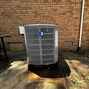 A gray American Standard air conditioning unit sits outdoors on a concrete pad in front of a red brick wall.