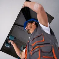 A worker in a blue hard hat and grey work vest uses a cordless drill to install a ceiling panel.