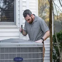 A technician inspects a Carrier HVAC unit outside a house using a handheld flashlight.