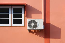 A white air conditioning unit mounted on a peach-colored wall next to a window with white frames.