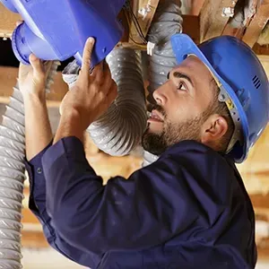 A person in a blue hard hat and uniform installs or repairs a blue ventilation component connected to a flexible duct.