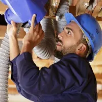 A person in a blue hard hat and uniform installs or repairs a blue ventilation component connected to a flexible duct.