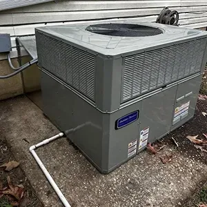 A gray American Standard air conditioning unit sits on a concrete pad next to a house exterior with light siding.