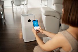 A person uses a smartphone to control a white air purifier in a modern, sunlit living room.