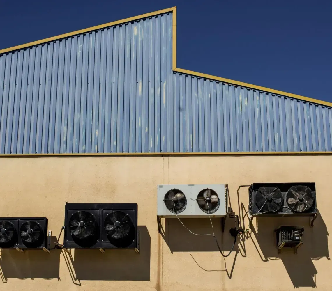 Four industrial air conditioning units mounted on a beige wall beneath a blue corrugated metal roof.