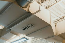A white rectangular HVAC supply duct suspended from a light-colored industrial or commercial ceiling.
