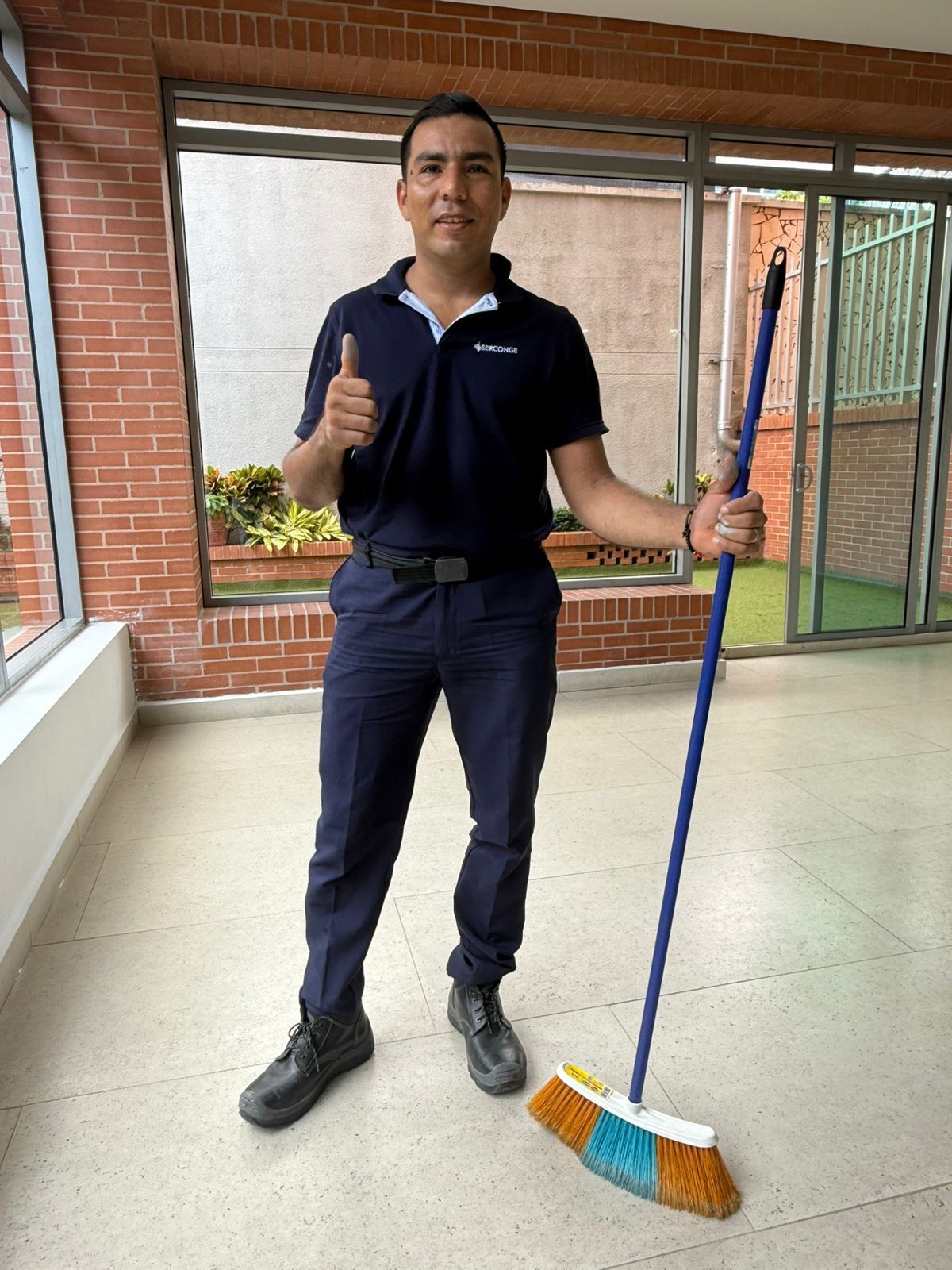Hombre uniformado con una escoba en la mano, haciendo el gesto de aprobación con el pulgar. Está de pie en el interior, junto a una ventana.
