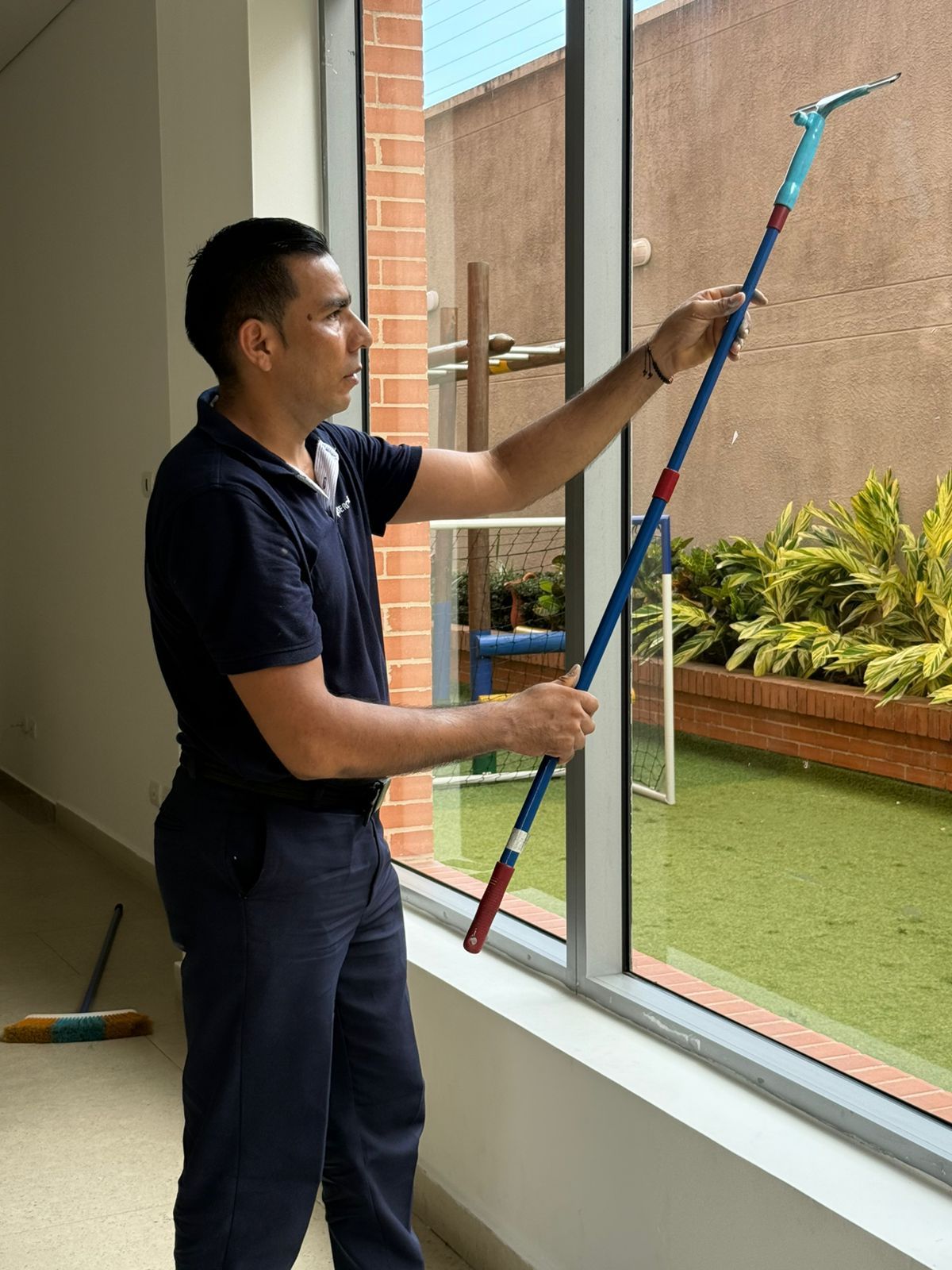 Hombre limpiando una ventana con un palo largo en un edificio, mirando hacia un patio.