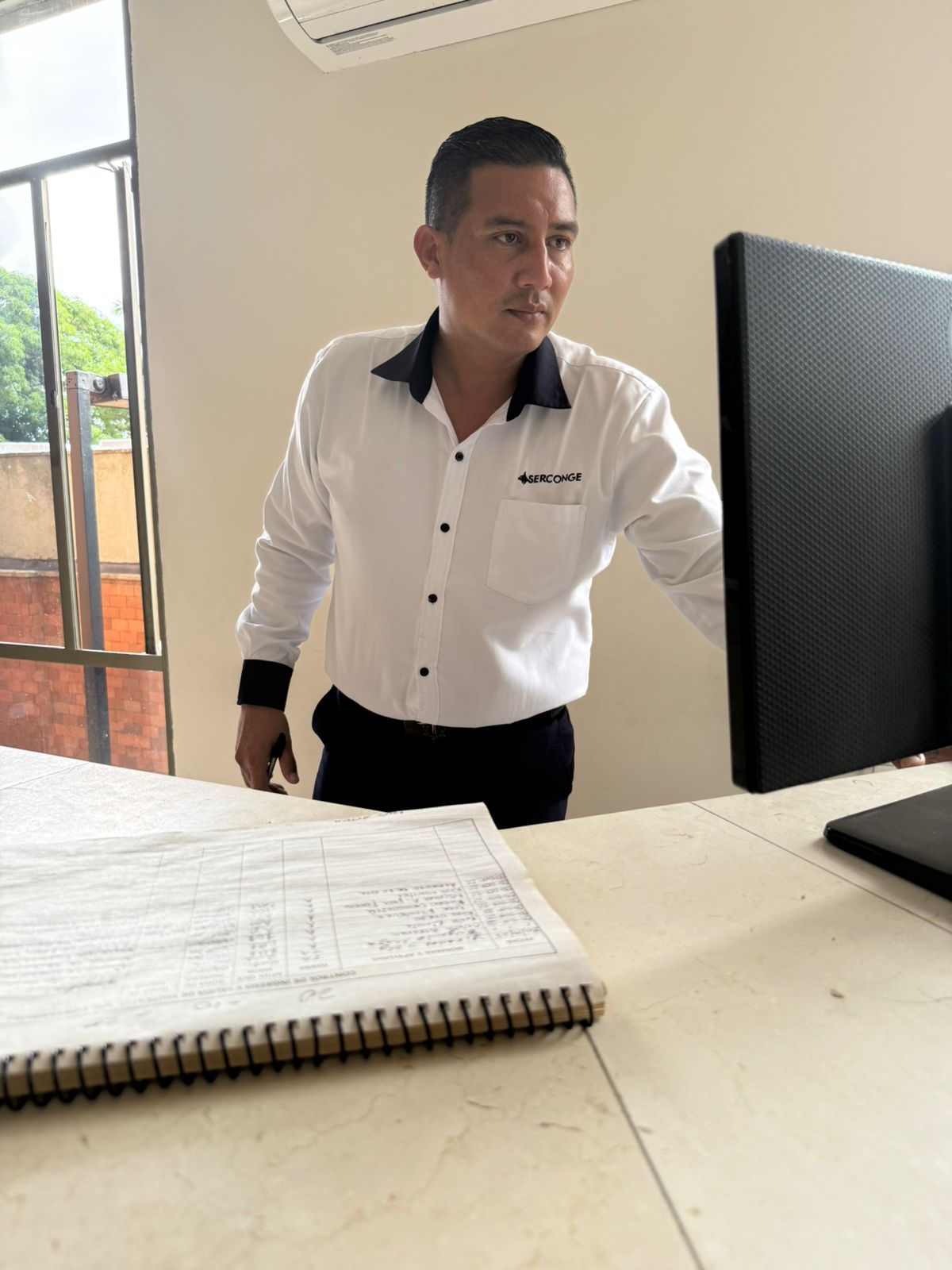 Hombre con camisa blanca y cuello negro mirando la pantalla de un ordenador, inclinado sobre un escritorio con un cuaderno.