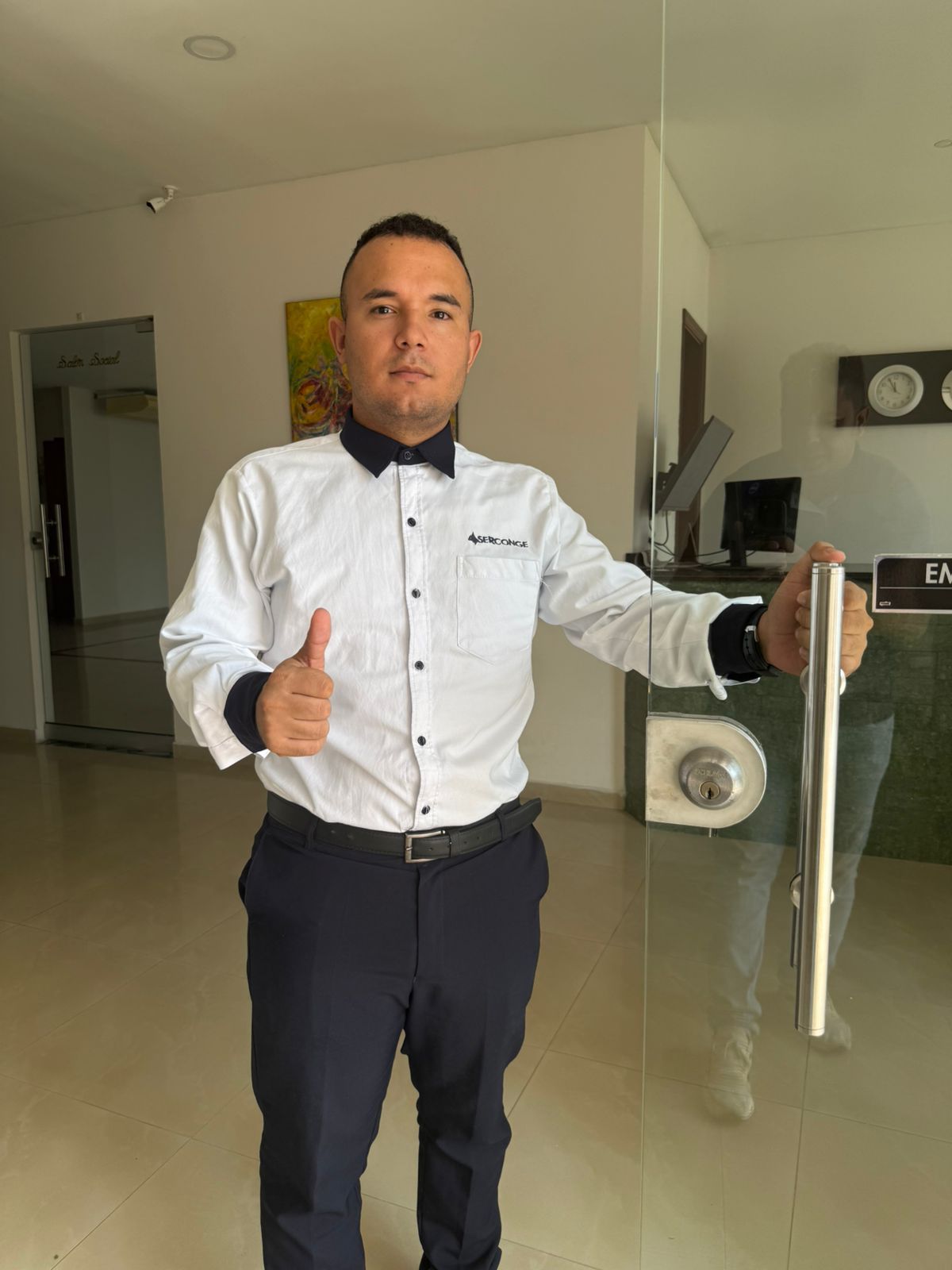 Hombre con camisa blanca y pantalones oscuros haciendo el gesto de aprobación con el pulgar en una oficina moderna, cerca de una puerta de cristal.