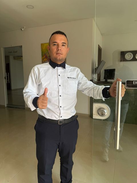 Hombre con camisa blanca y pantalones oscuros haciendo el gesto de aprobación con el pulgar en una oficina moderna, cerca de una puerta de cristal.