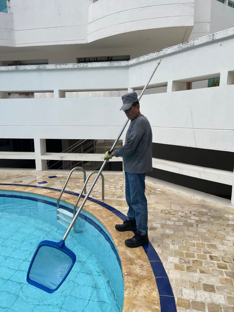 Persona limpiando una piscina azul con una red, cerca de un edificio blanco.