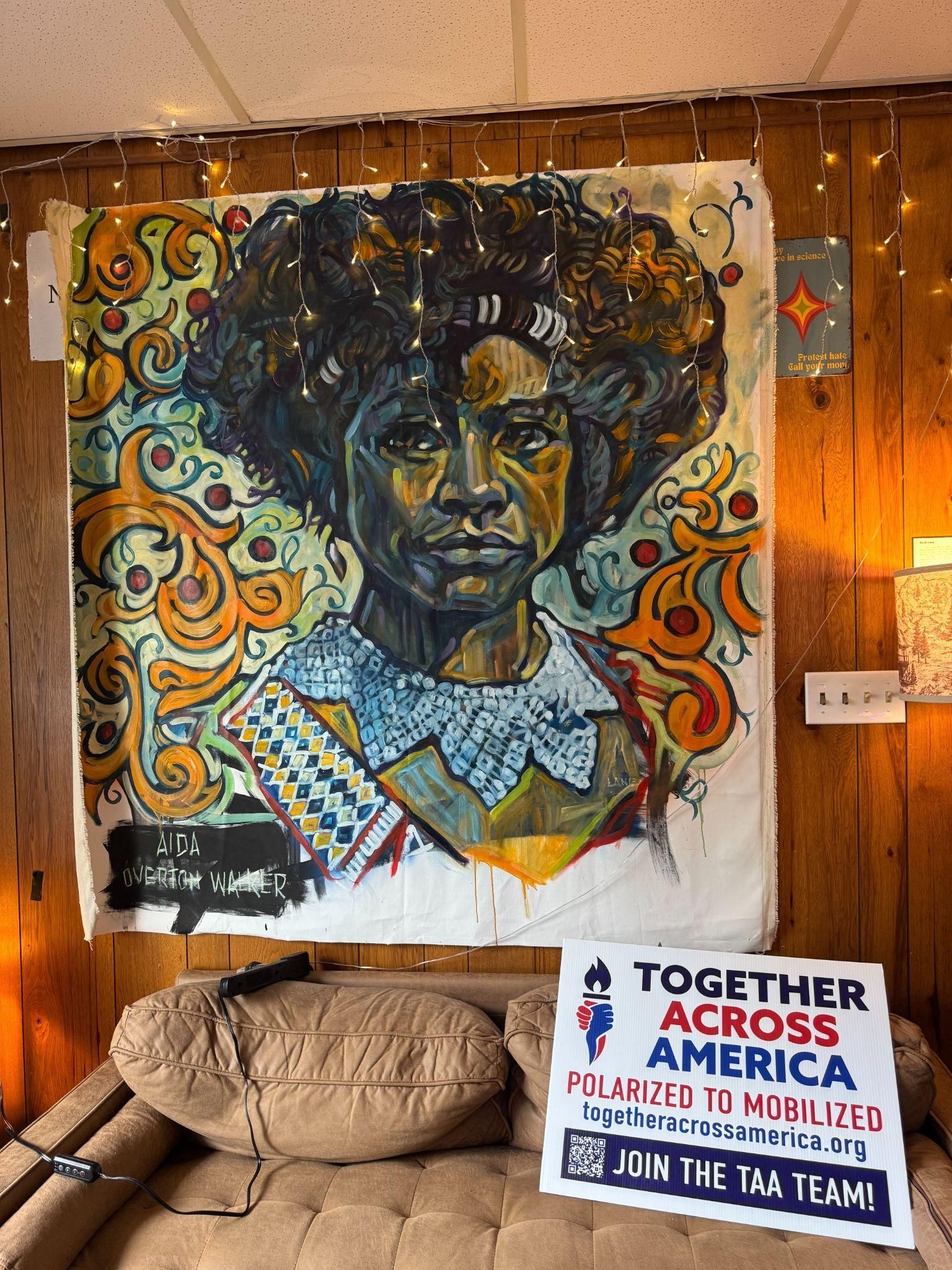 A large, colorful portrait of a person hangs above a couch next to a sign that reads