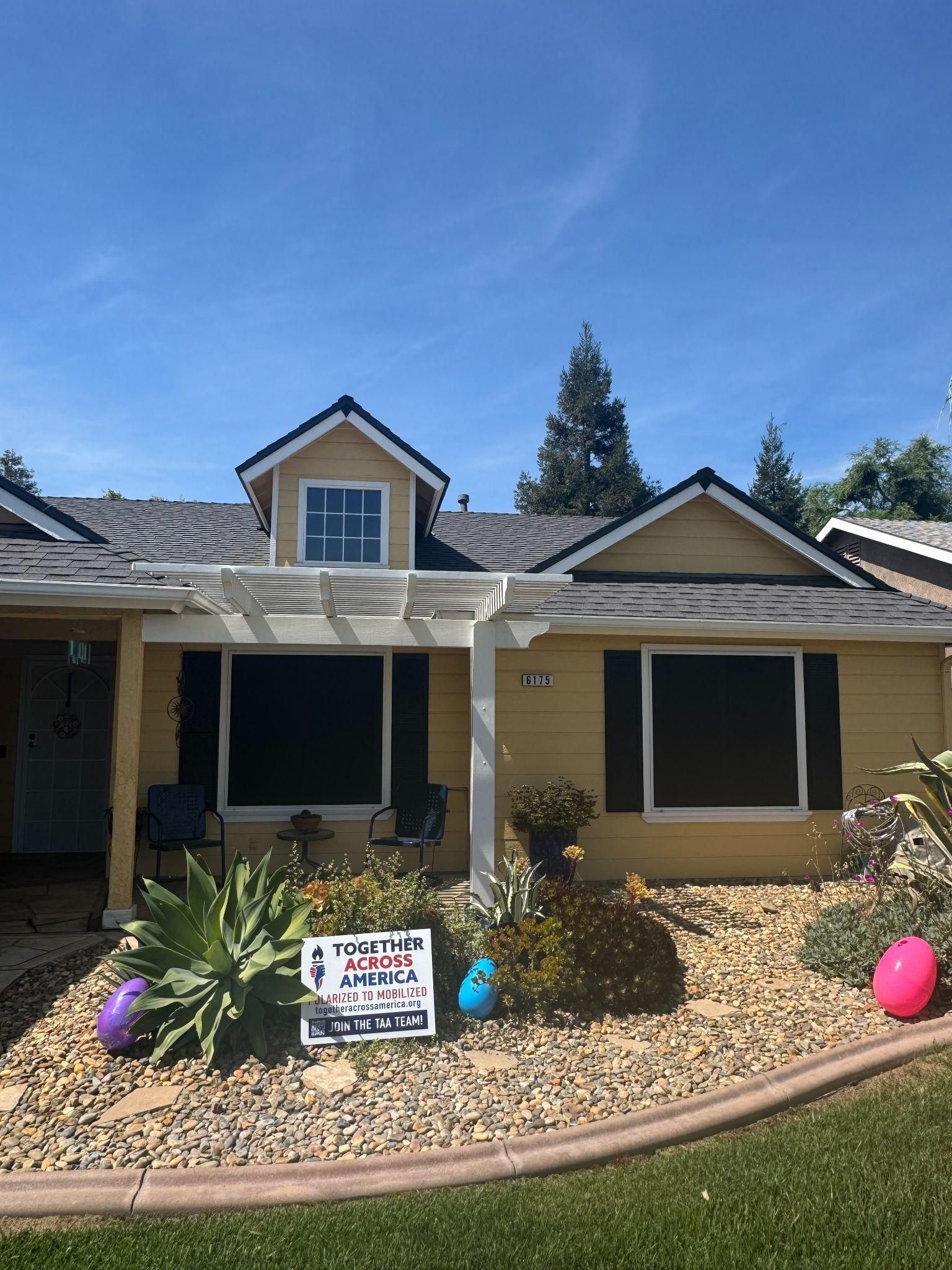 A single-story, yellow house with dark shutters and a white pergola, set against a bright blue sky with Easter decorations.