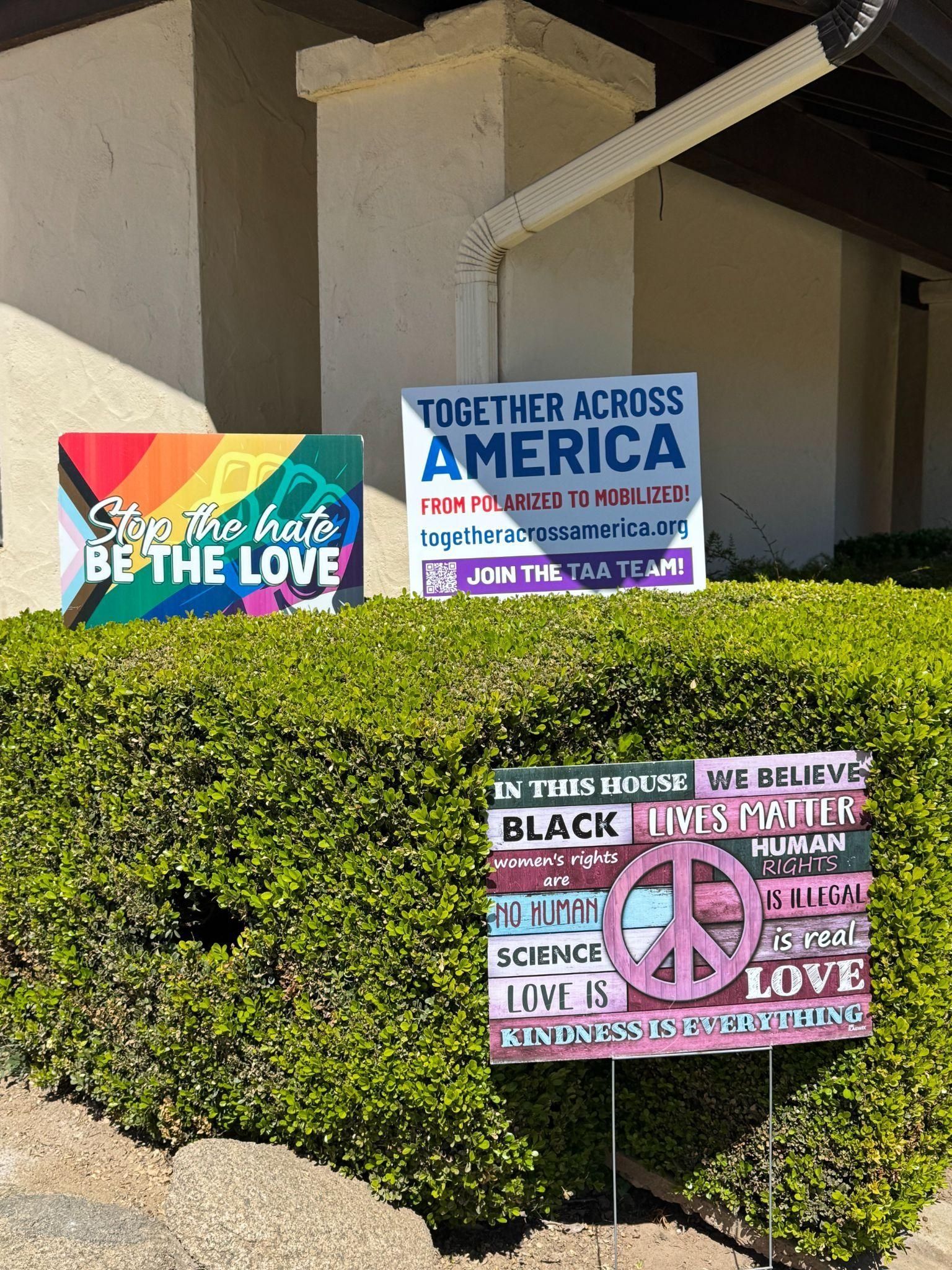 Three signs with messages of inclusion, social justice, and peace are displayed in front of a green hedge.