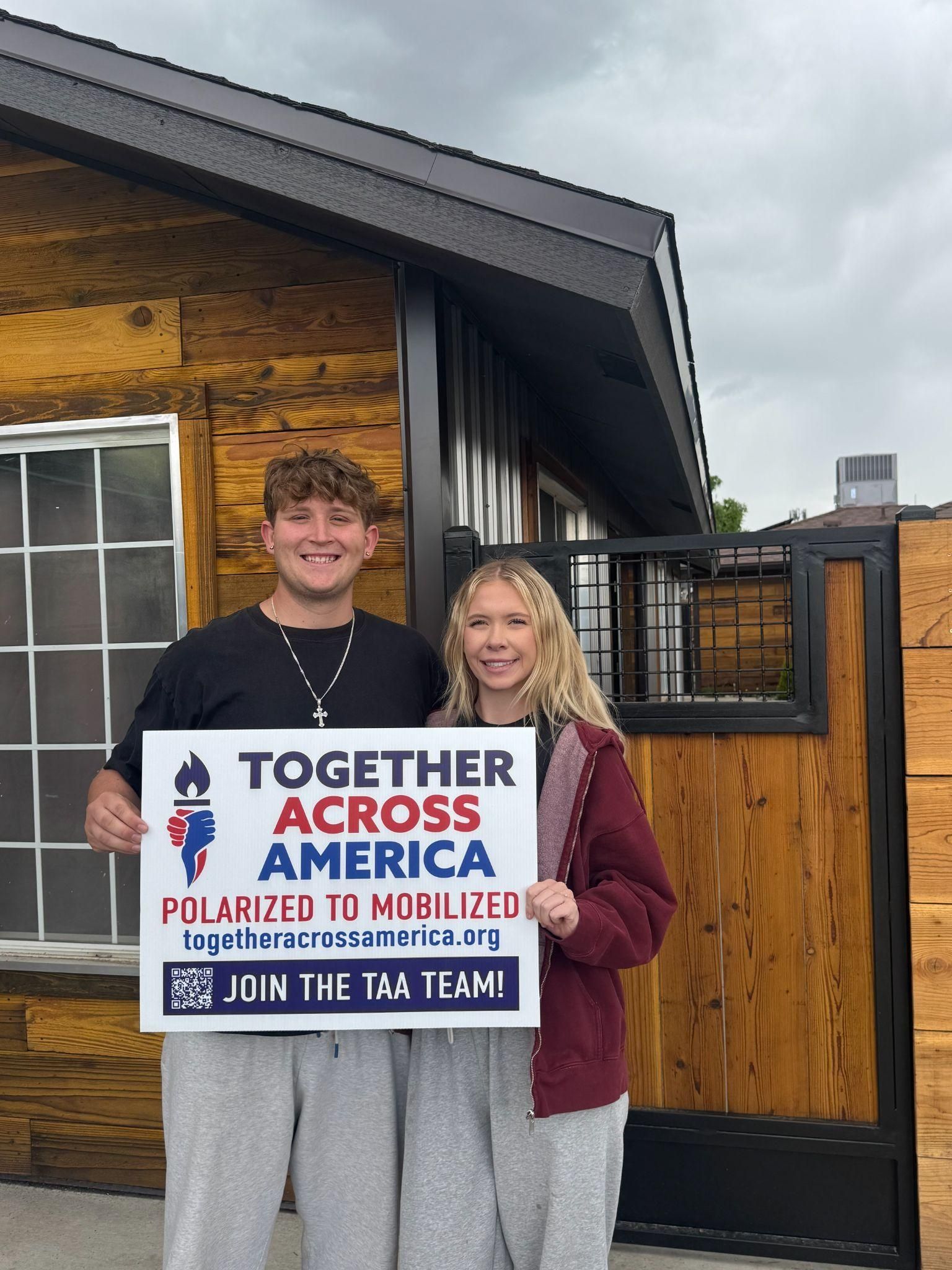Two people stand in front of a wood-paneled building, holding a sign that reads