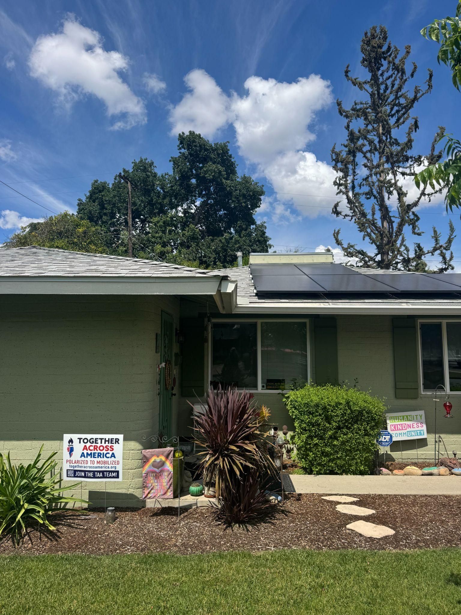 A light green house with solar panels on the roof, surrounded by small bushes, a yard sign, and a tall tree under a blue sky.