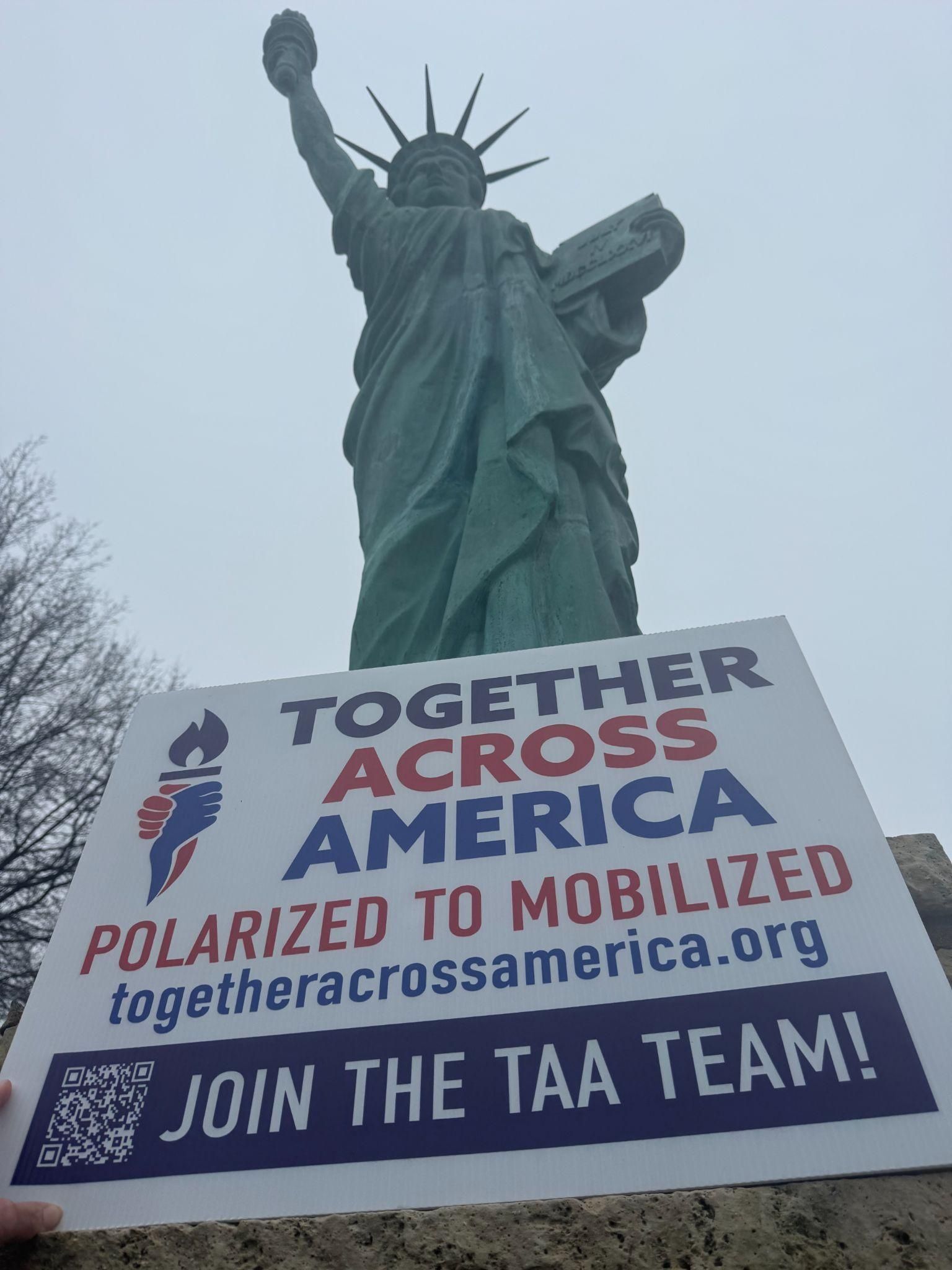A hand holds a sign in front of the Statue of Liberty that says