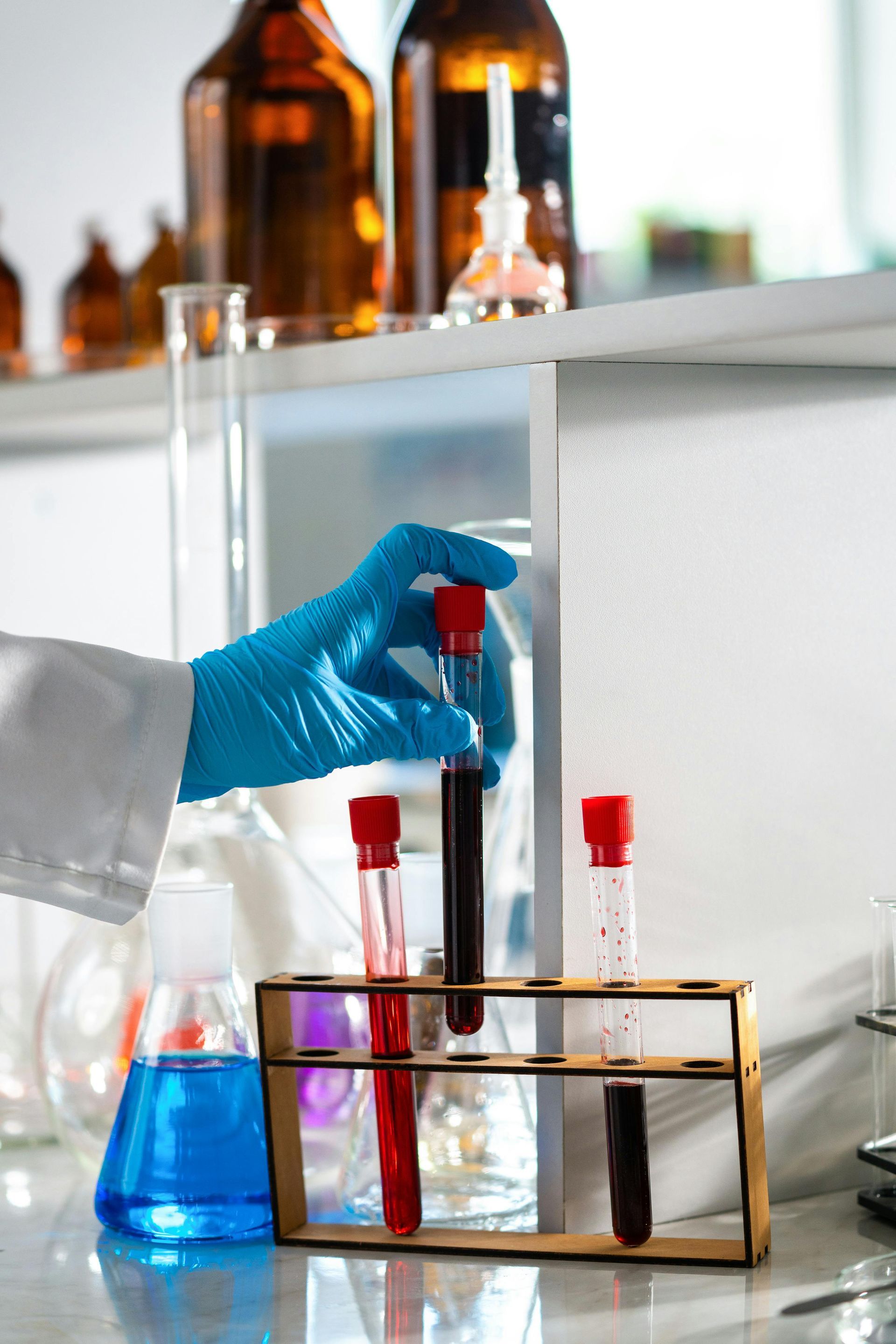 Gloved hand holding a test tube in a lab with glassware and red and blue liquids