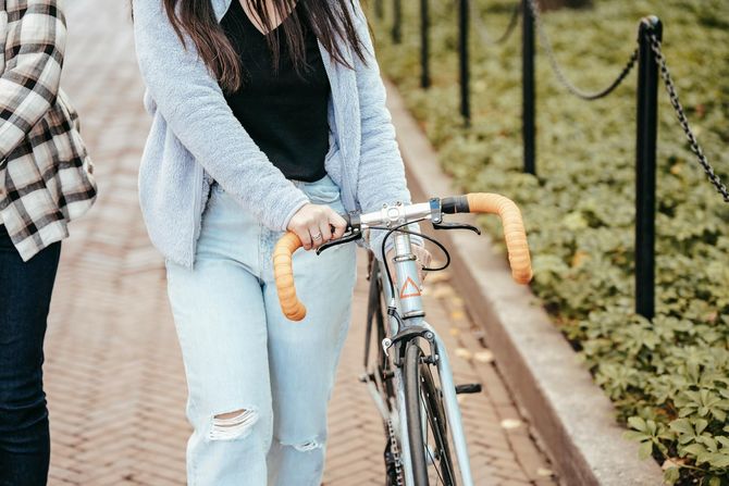 Person holding a silver bicycle with orange handlebars. They wear light blue jeans and a gray cardigan.