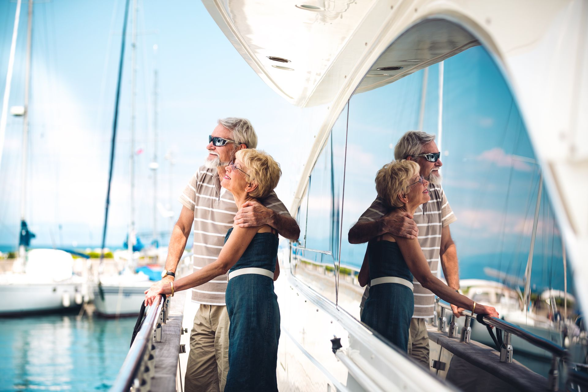 Couple on a yacht looking at sailboats; reflections in the boat's window. Sunny day, water.