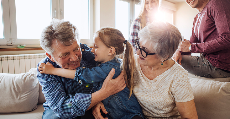 Grandparents embracing a young child on a couch, other family members in the background.