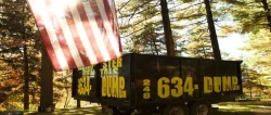 American flag hanging beside a black-and-yellow 634-DUMP dumpster in a wooded area