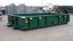 Green industrial dumpster or roll-off container on a paved lot with trucks in the background