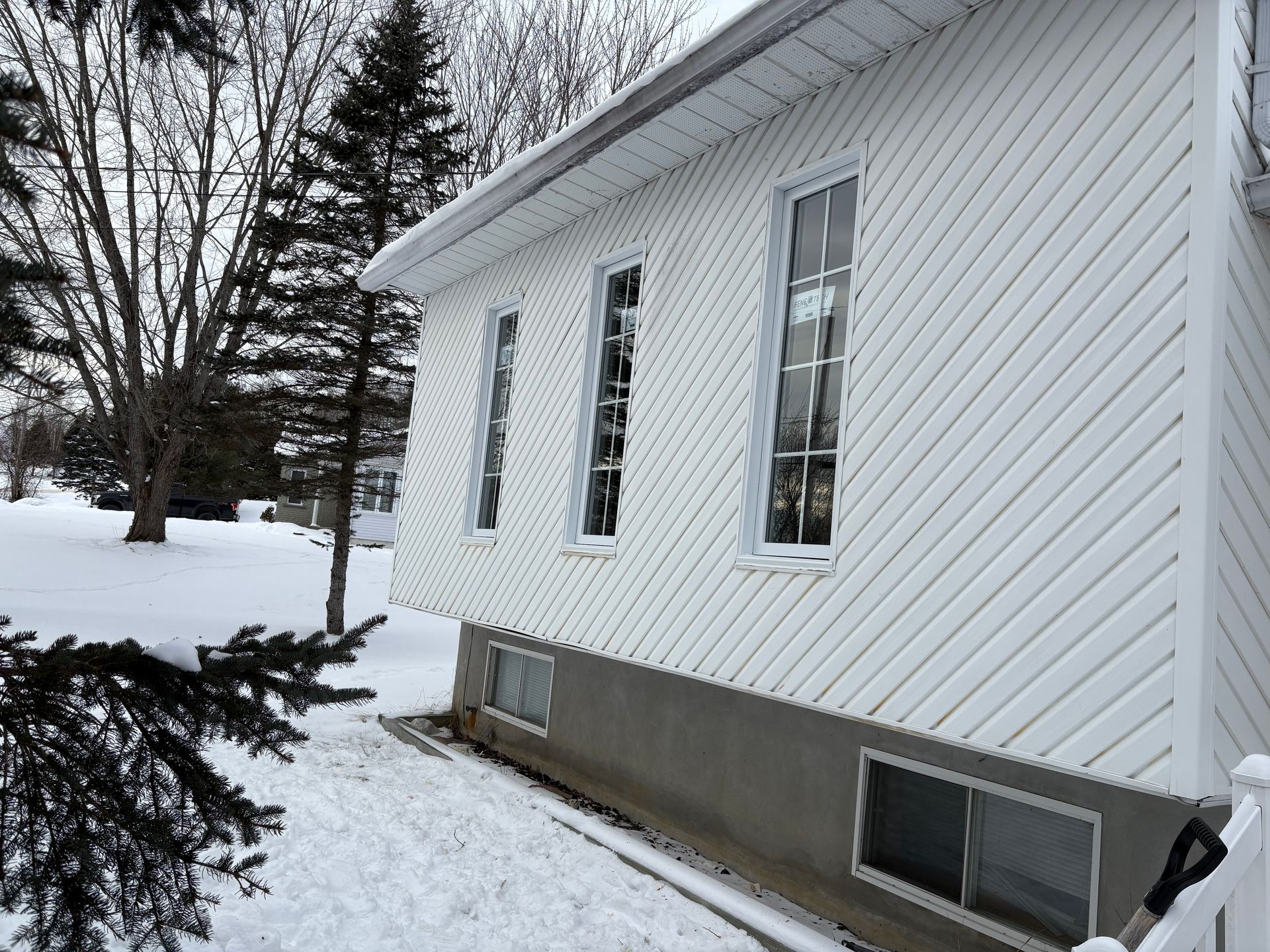 Façade d'une maison blanche recouverte de neige, avec des fenêtres au sous-sol et des arbres à proximité