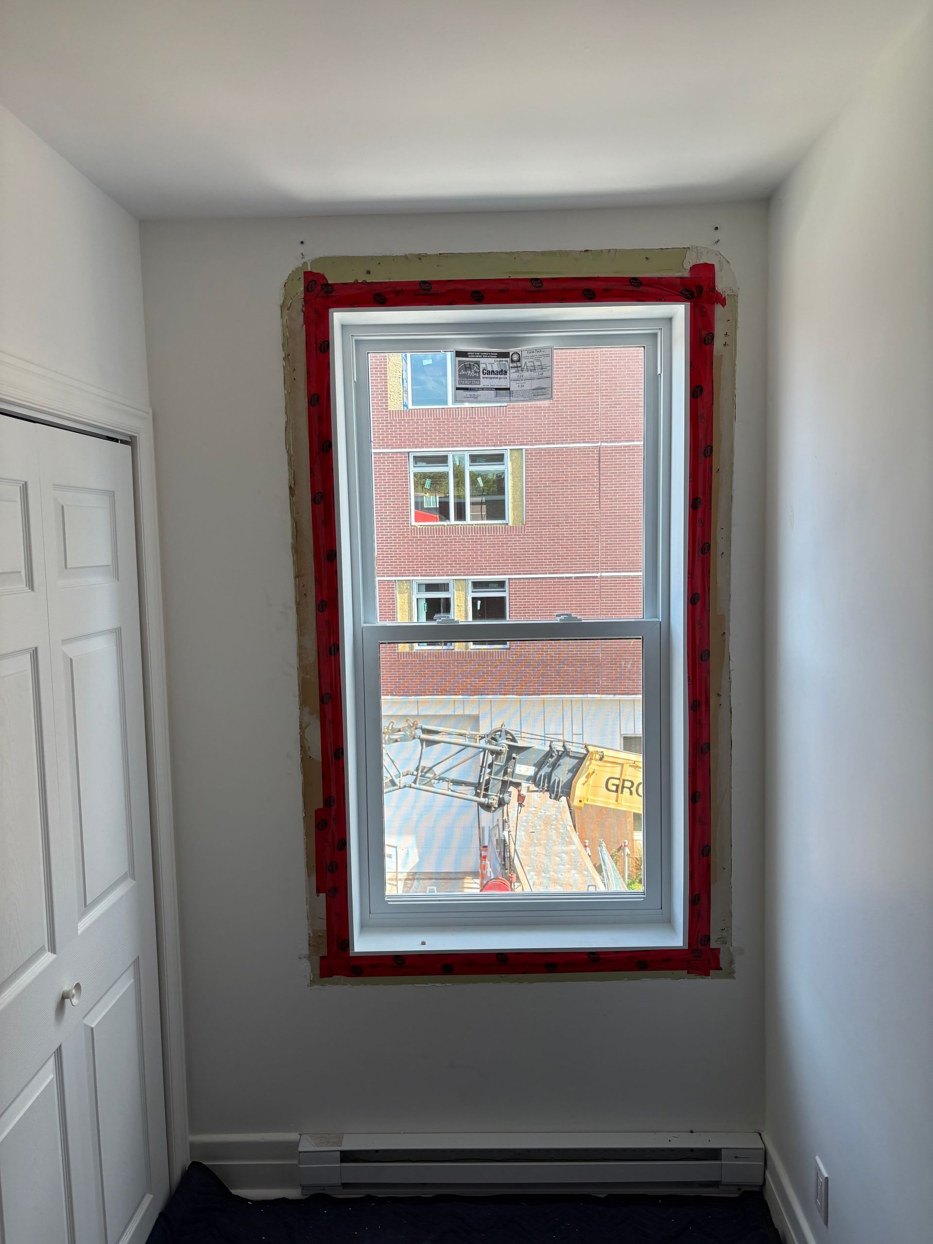 Pièce blanche vide avec une unique fenêtre encadrée de rouge, donnant sur un bâtiment en briques.