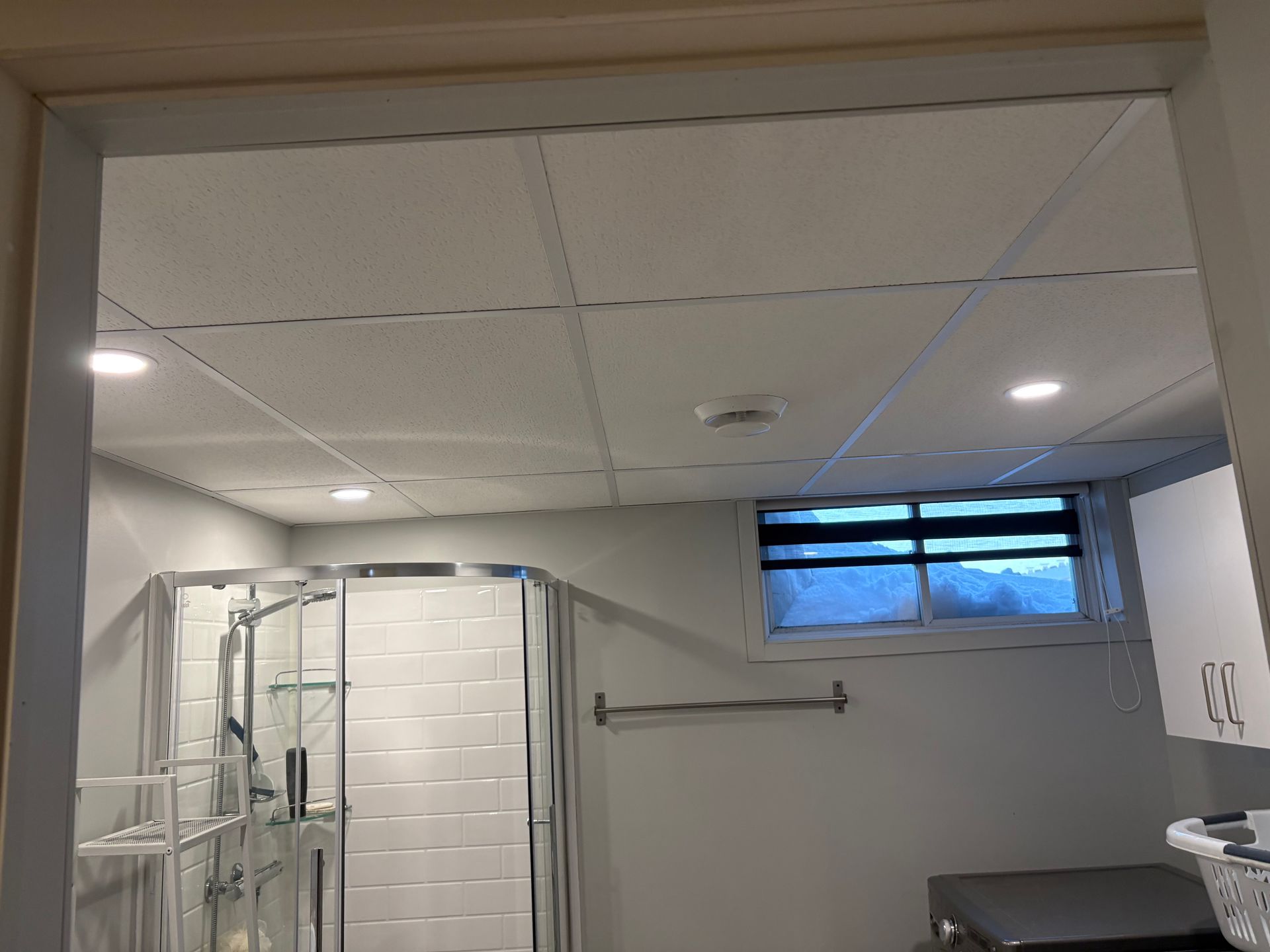Salle de bains avec douche carrelée, plafond en carreaux blancs et petite fenêtre dépolie au-dessus d'un plan de travail.