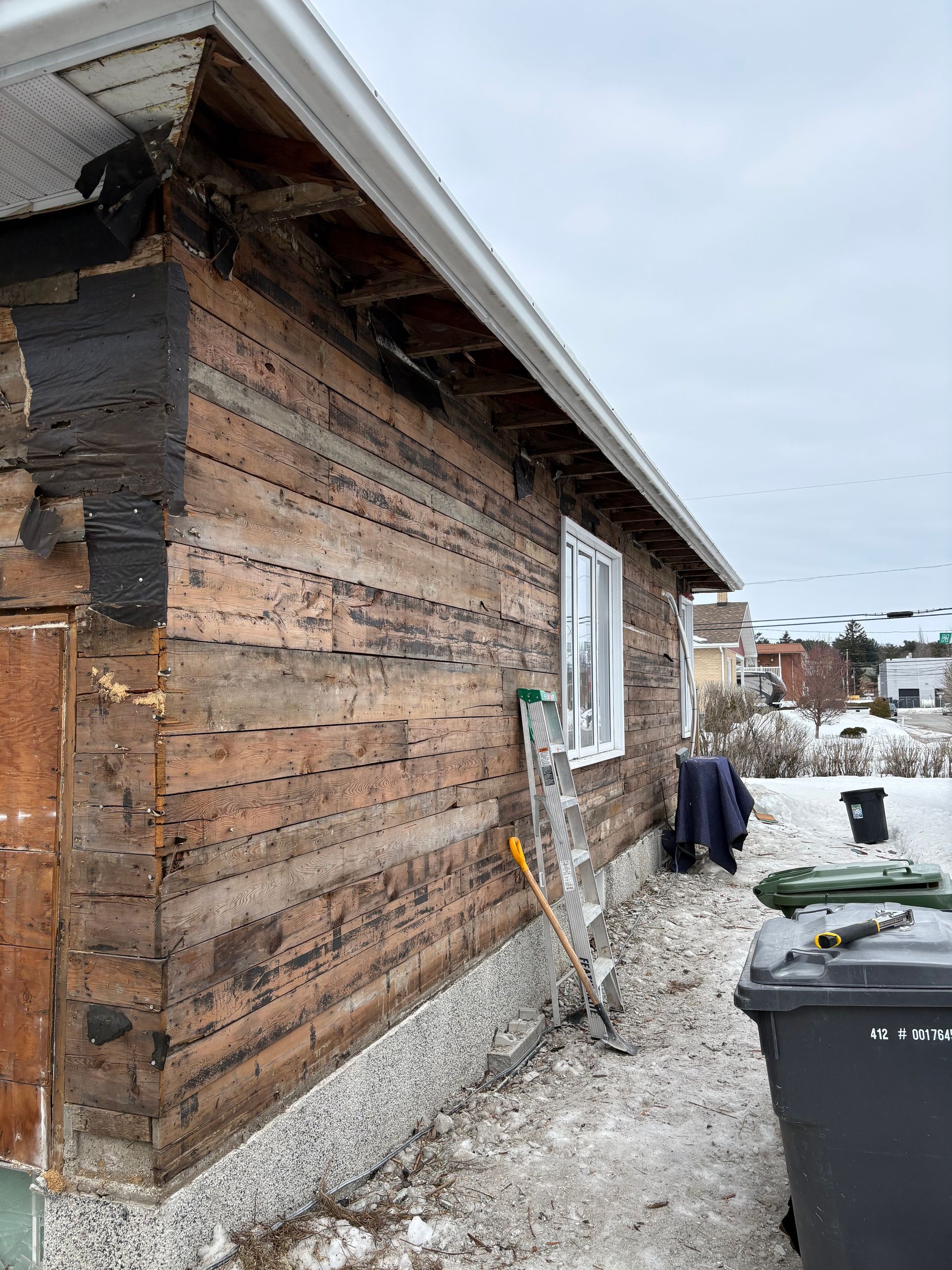 Abri de jardin en bois délabré, au toit enneigé, à côté de poubelles dans une cour en gravier