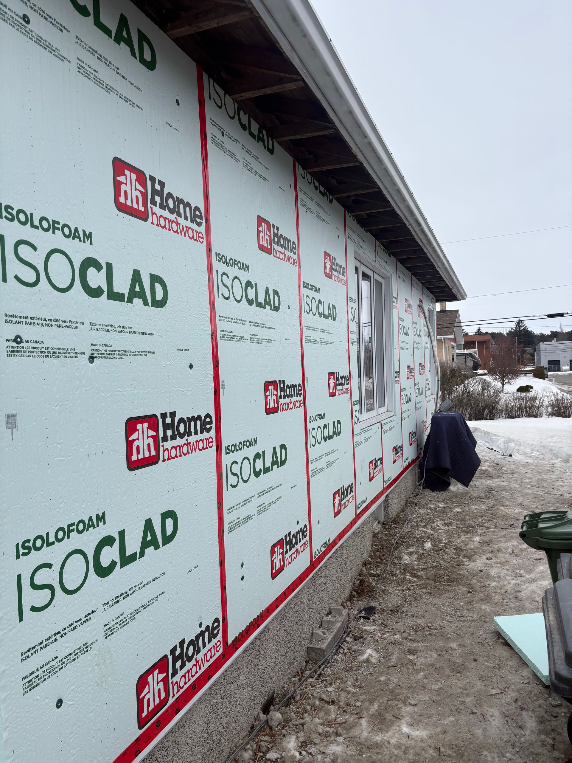 Mur extérieur en construction, recouvert d'isolant blanc ISOCLAD, avec un ouvrier accroupi près du sol enneigé.