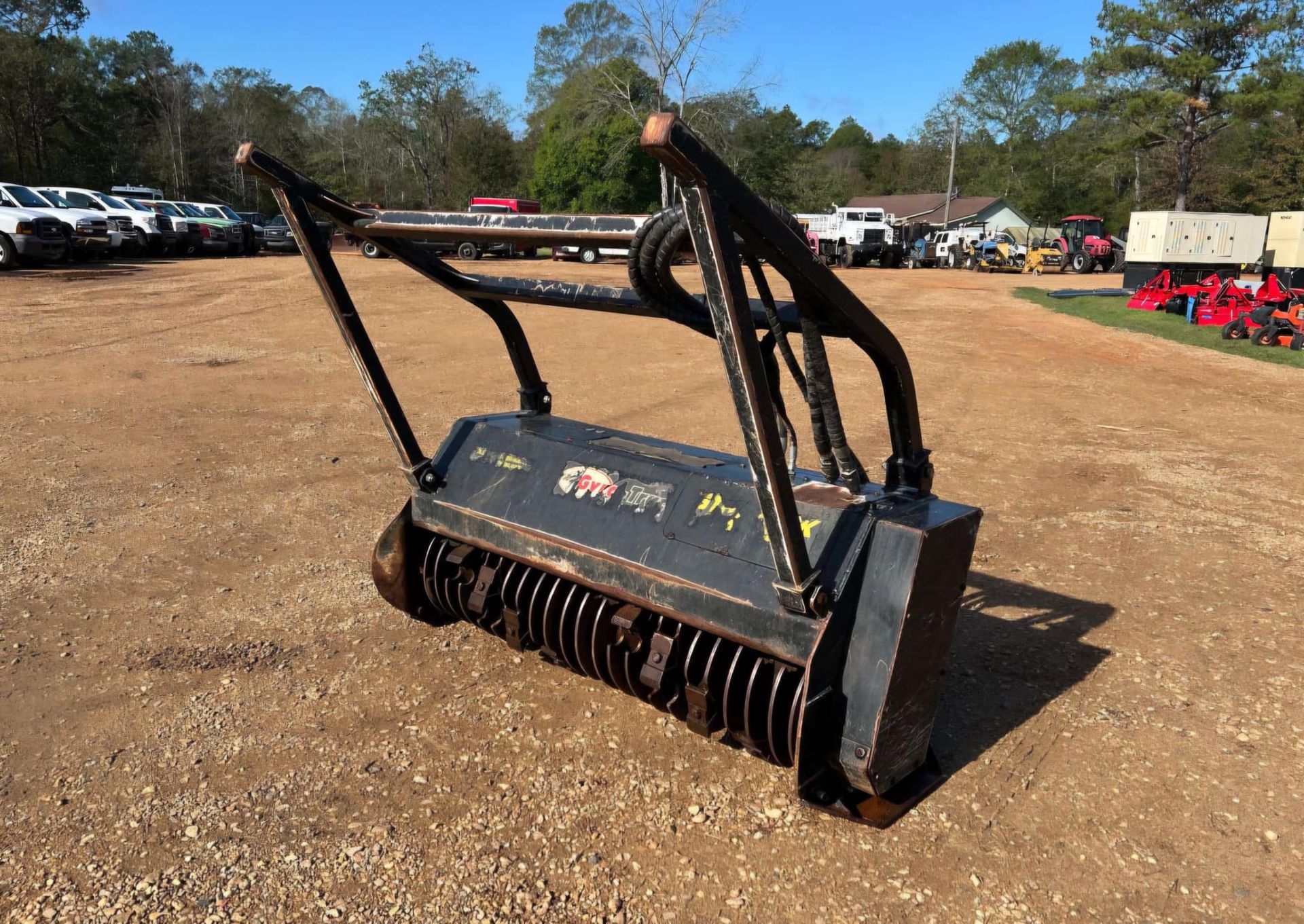 Brush cutter attachment, black and rust-colored metal, on gravel lot, with vehicles in the background.